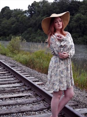 A figure in a floral dress and sun hat poses along rustic train tracks in a natural outdoor setting.
