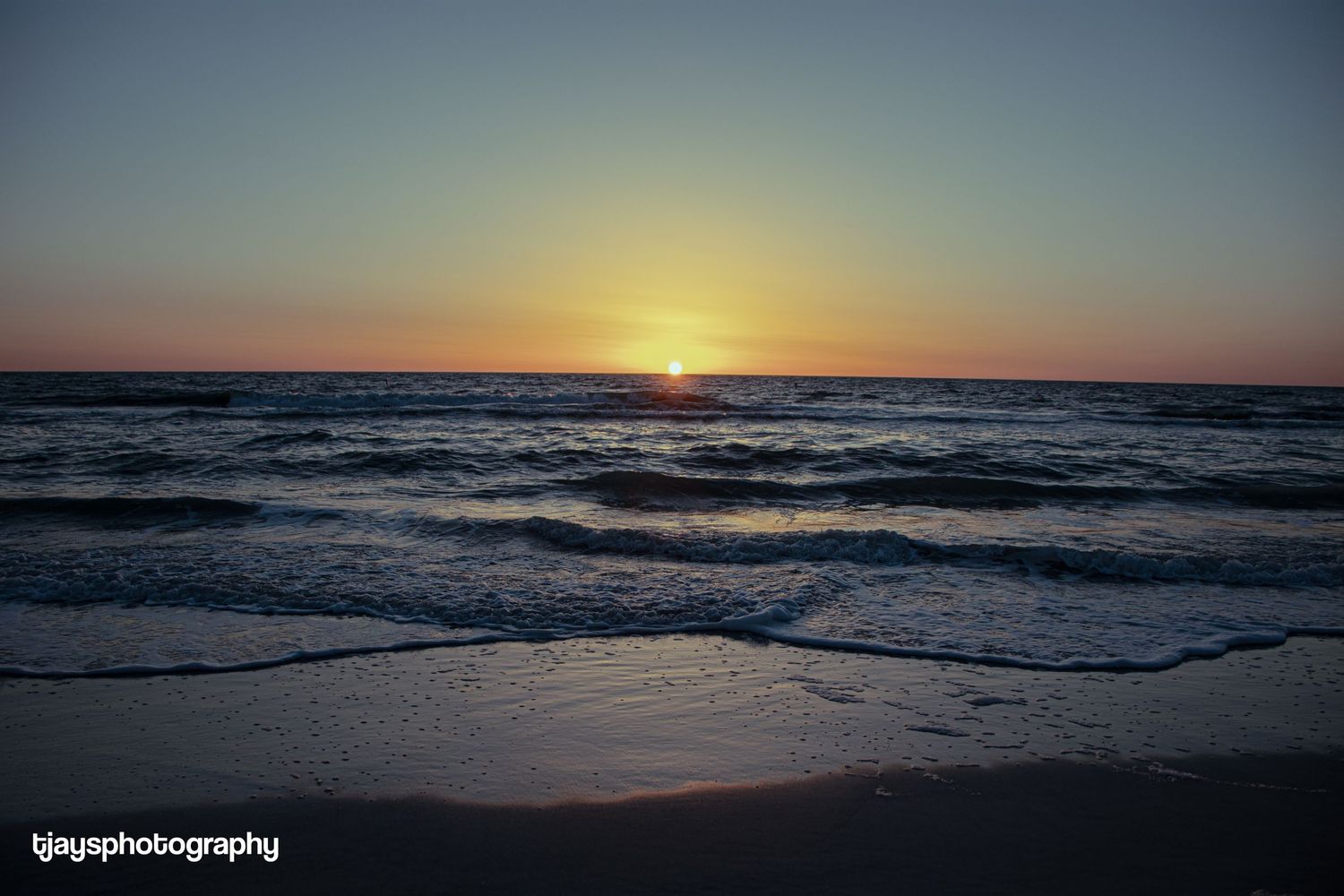 A stunning ocean sunset with golden sun touching the horizon over gentle rolling waves on the beach.