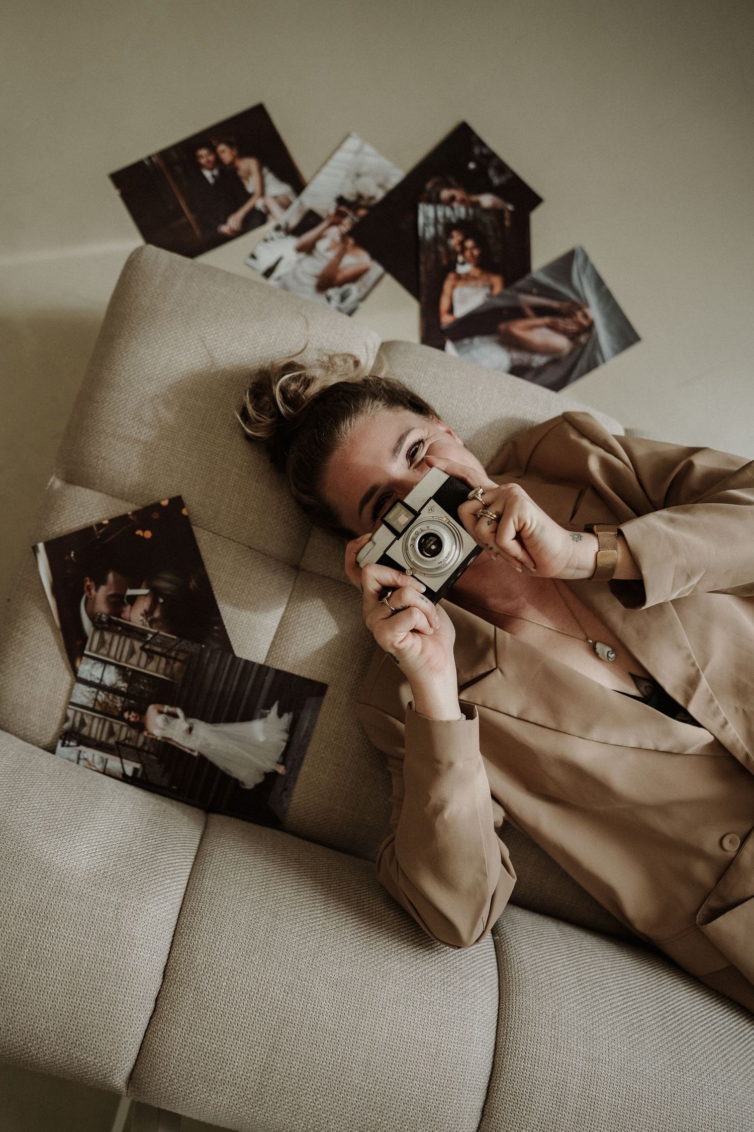 A vintage camera held up while lying on a beige couch with scattered photographs in the background.
