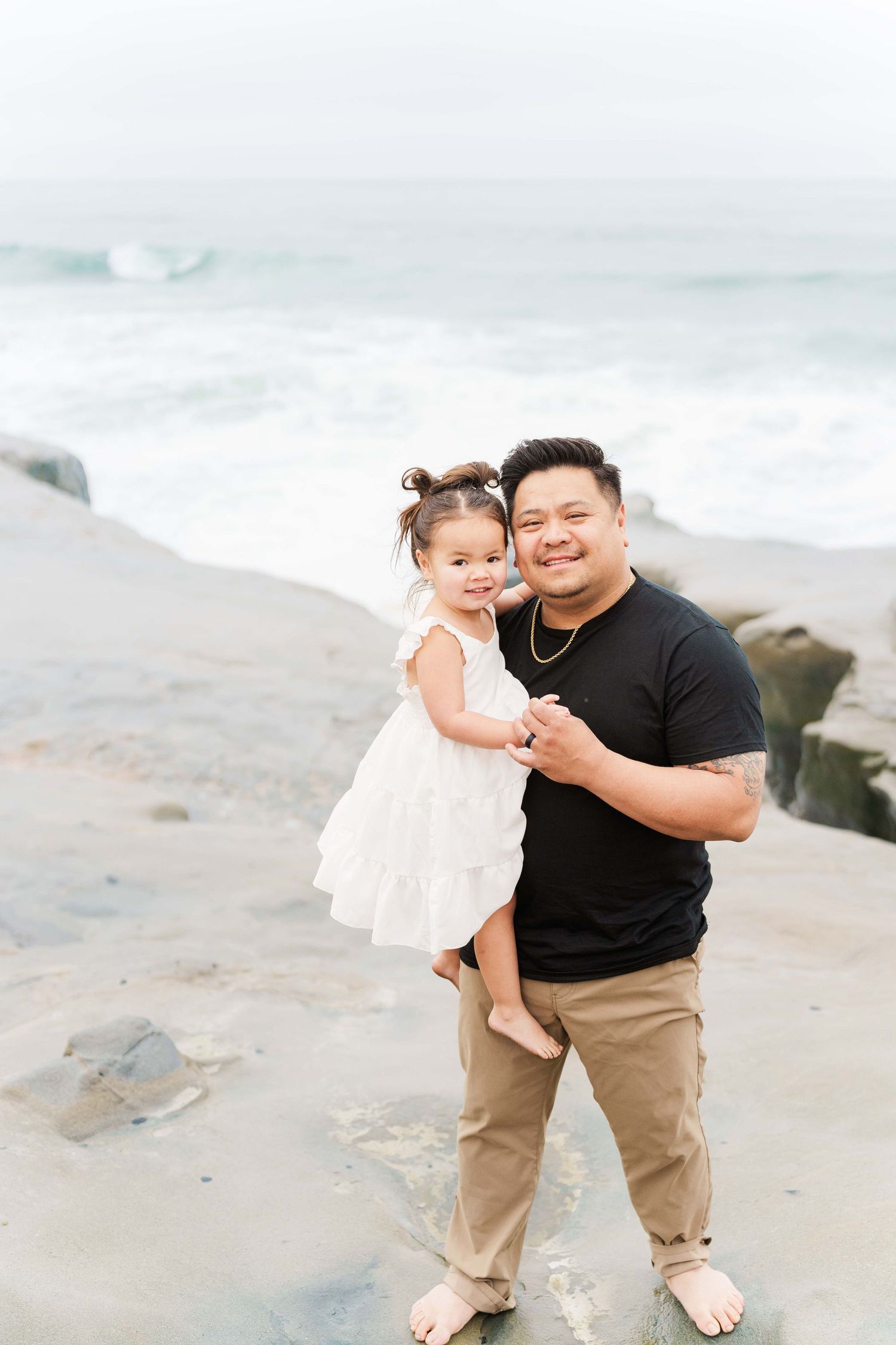 A heartwarming moment on a misty beach with waves crashing in the background as they stand on rocky sand.