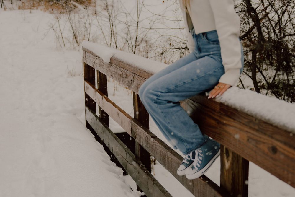 Person in blue jeans and white sneakers sits on a snow-covered wooden railing during winter.