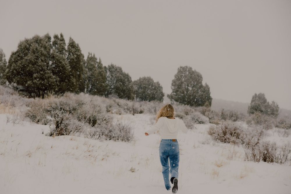 A person in denim jeans walks through a snowy winter landscape with trees in the background.