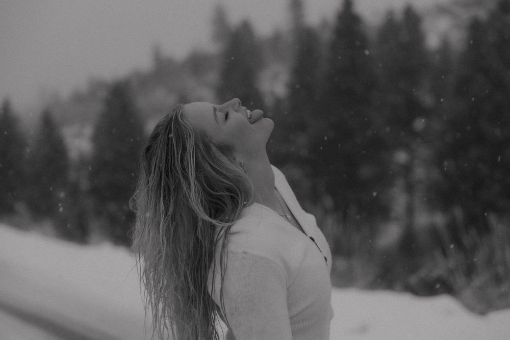 A joyful figure in a white sweater tilts their head back to catch falling snow against a wintery forest backdrop.