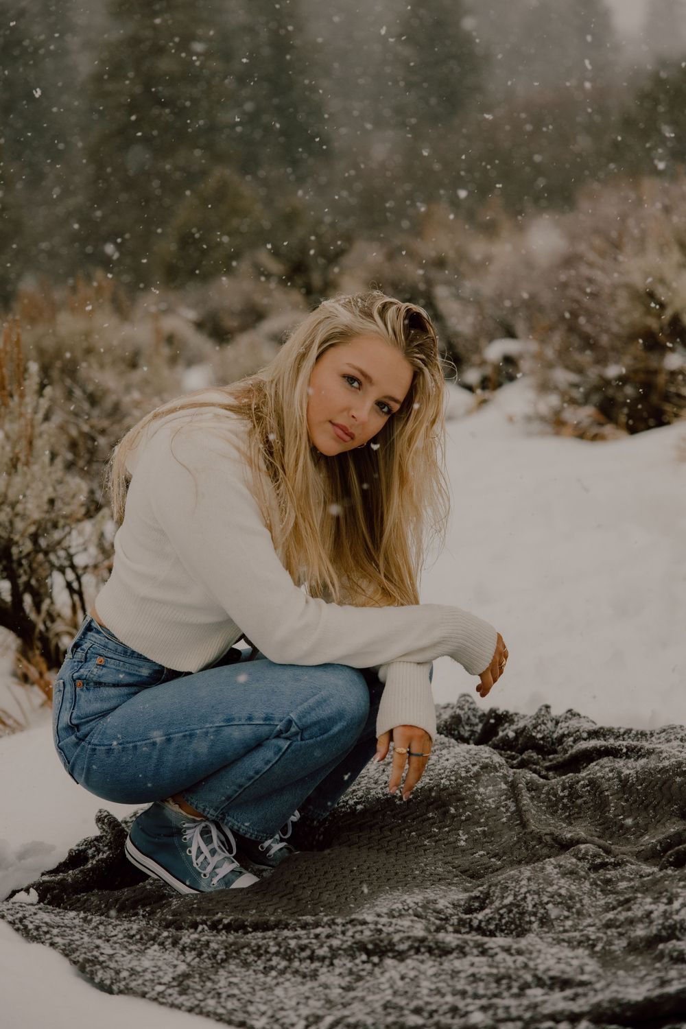 A person in a white sweater and blue jeans crouches in the snow during a winter photoshoot.