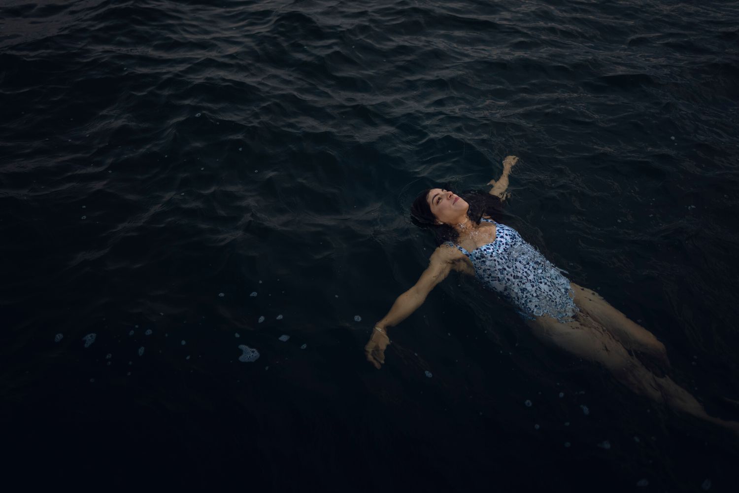 A lady swims on her back diagonally across dark lake water in Lake Ontario Park, Kingston, Ontario.