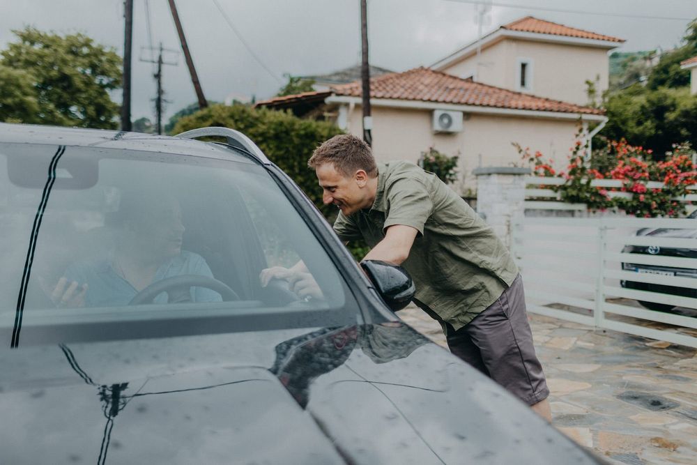 Someone leans over to inspect a black car in a residential driveway on a cloudy day.