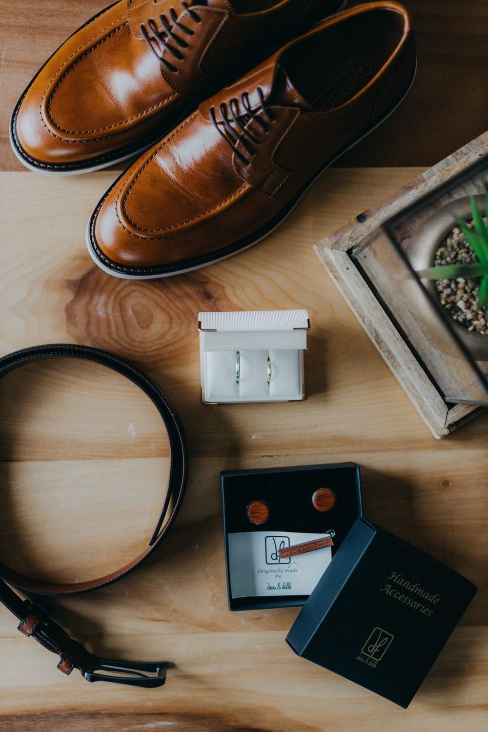 Brown leather dress shoes displayed with accessories including a belt, passport and cufflinks on wooden surface.