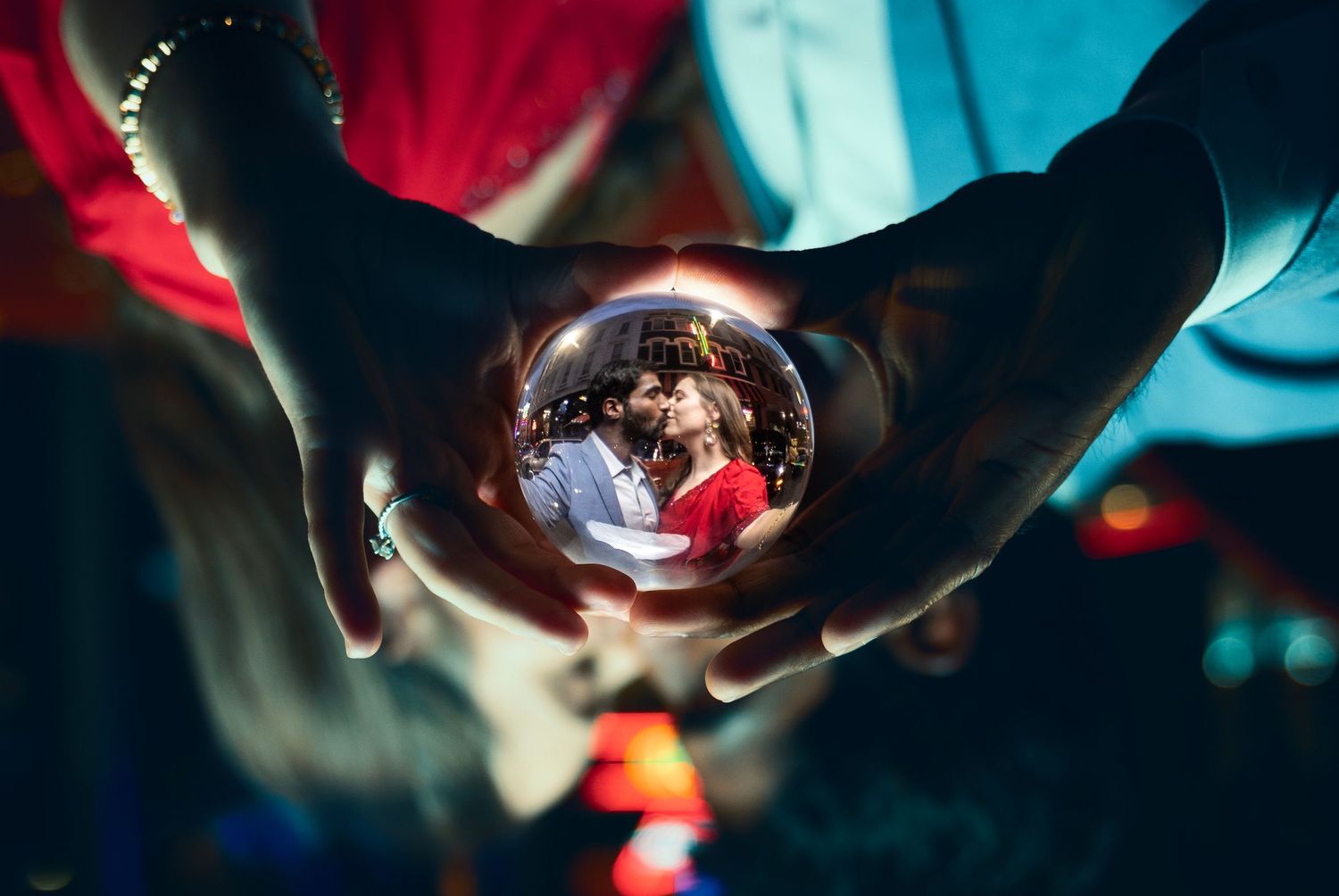 Two hands hold a glass crystal ball reflecting a romantic couple in an embrace, with colorful bokeh lights glowing in the dark background.
