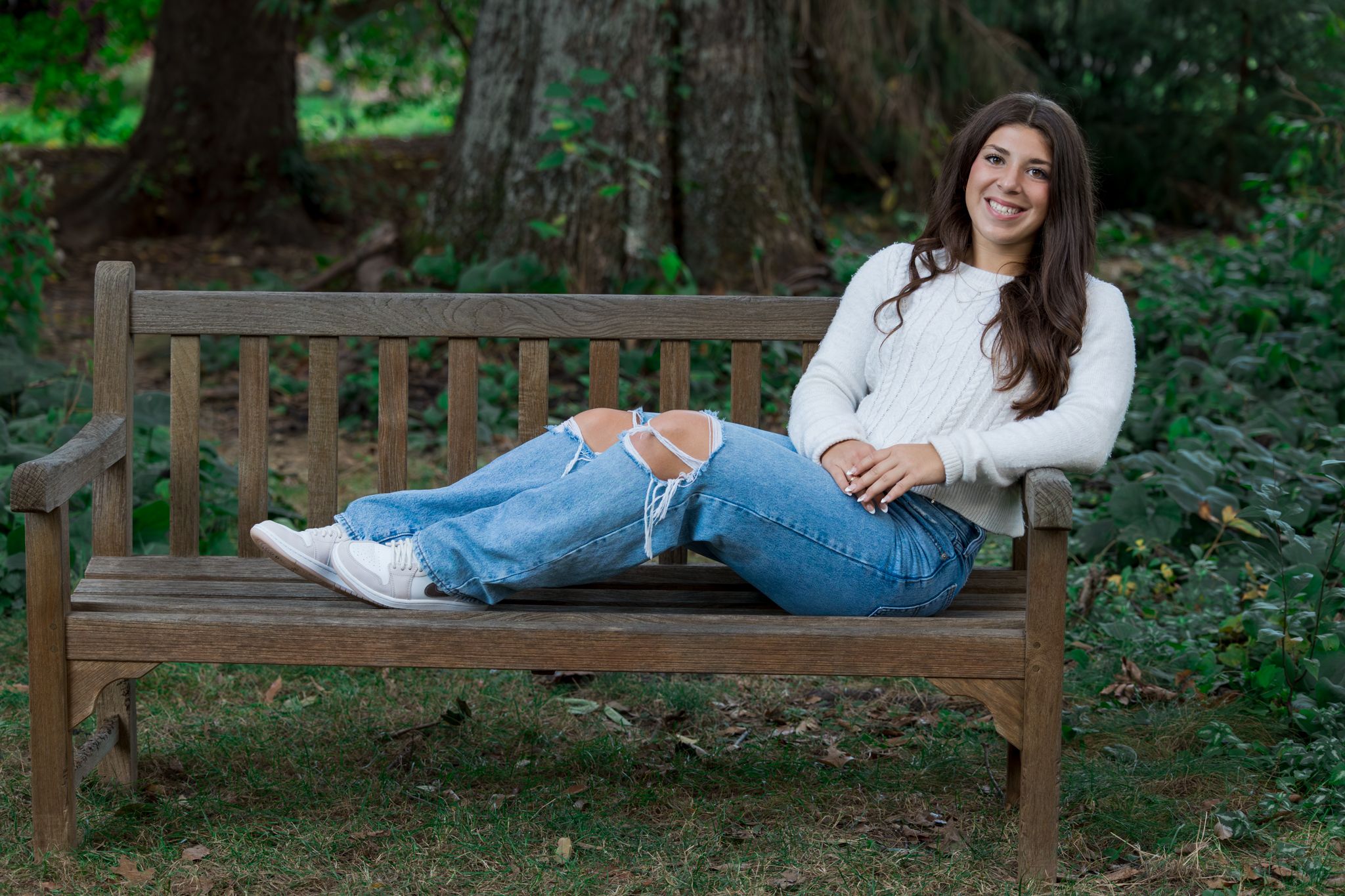 Person in casual attire lounges on wooden bench in lush garden setting, surrounded by green foliage.