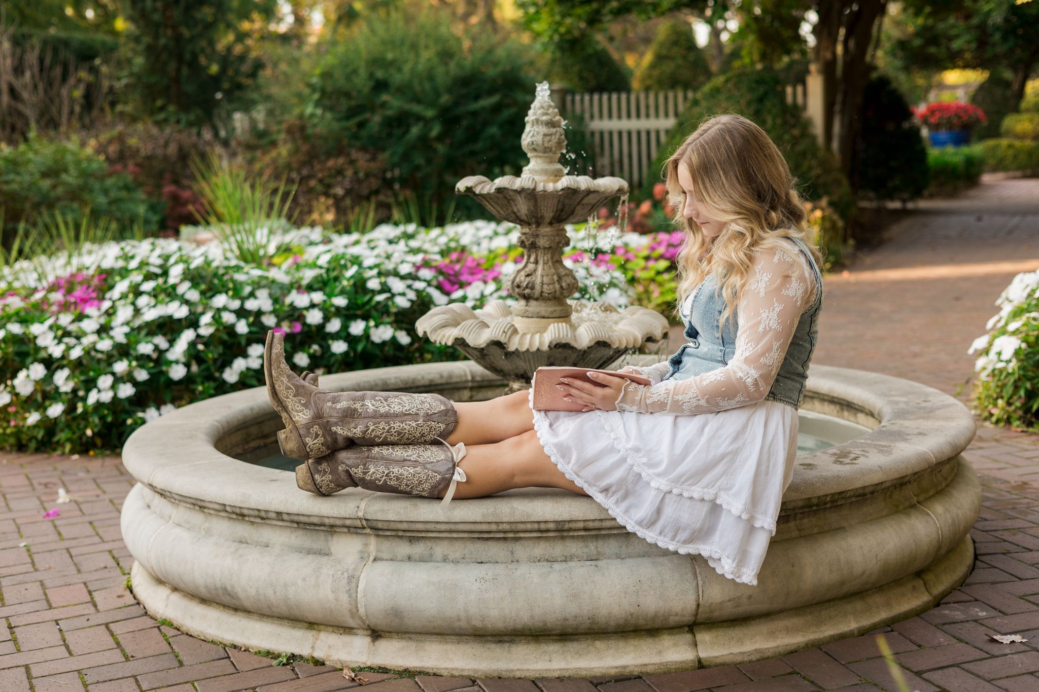 A person in a white dress sits on the edge of a decorative stone fountain in a lush garden setting.