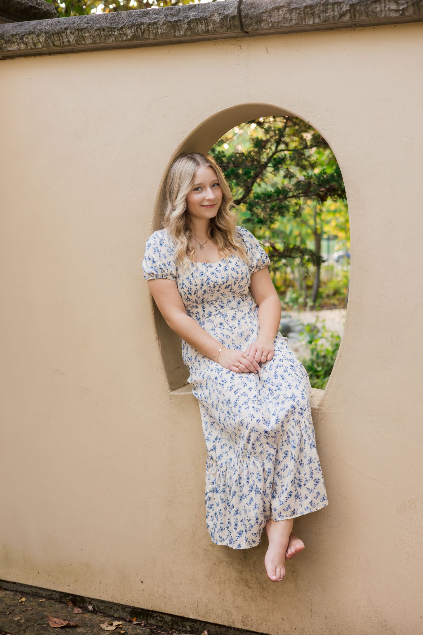 A person in a white floral dress sits gracefully on a beige wall in an outdoor garden setting.