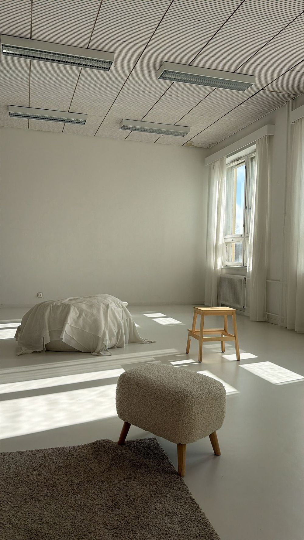 Bright minimalist white room with a bed on the floor, wooden stool, and natural sunlight.