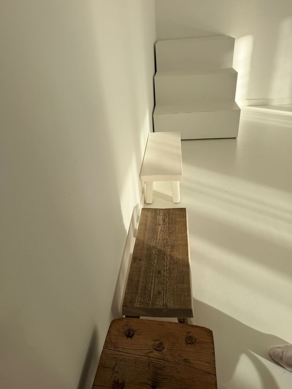 Sunlit white staircase with a rustic wooden step stool casting shadows on the wall.