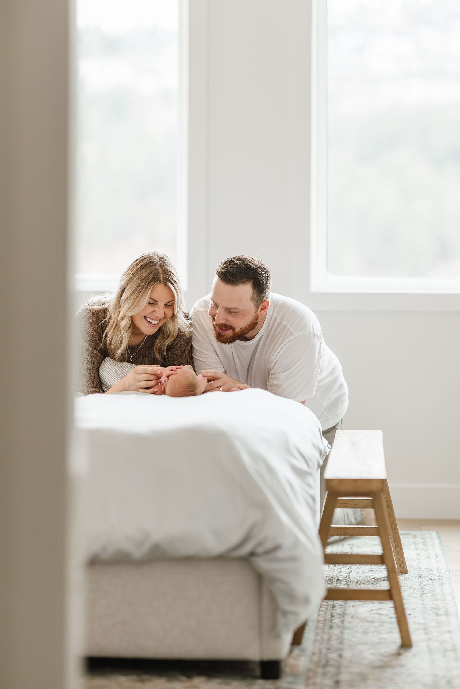 Couple lying on white bedding in bright minimalist bedroom with wooden chair creating intimate lifestyle moment.