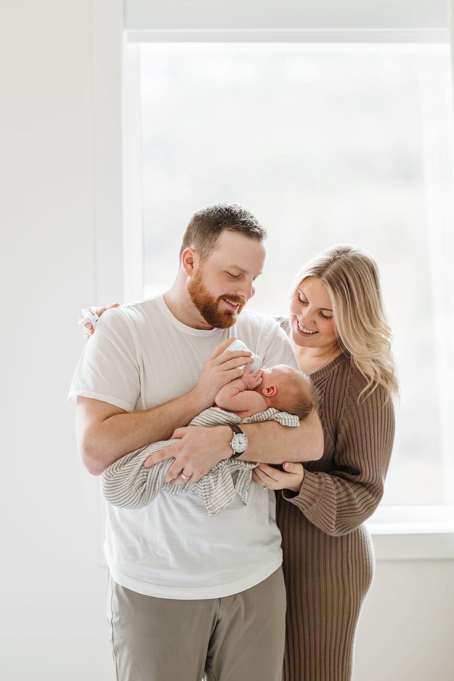 A couple in casual attire shares joyful moments with their newborn baby in a bright white room.
