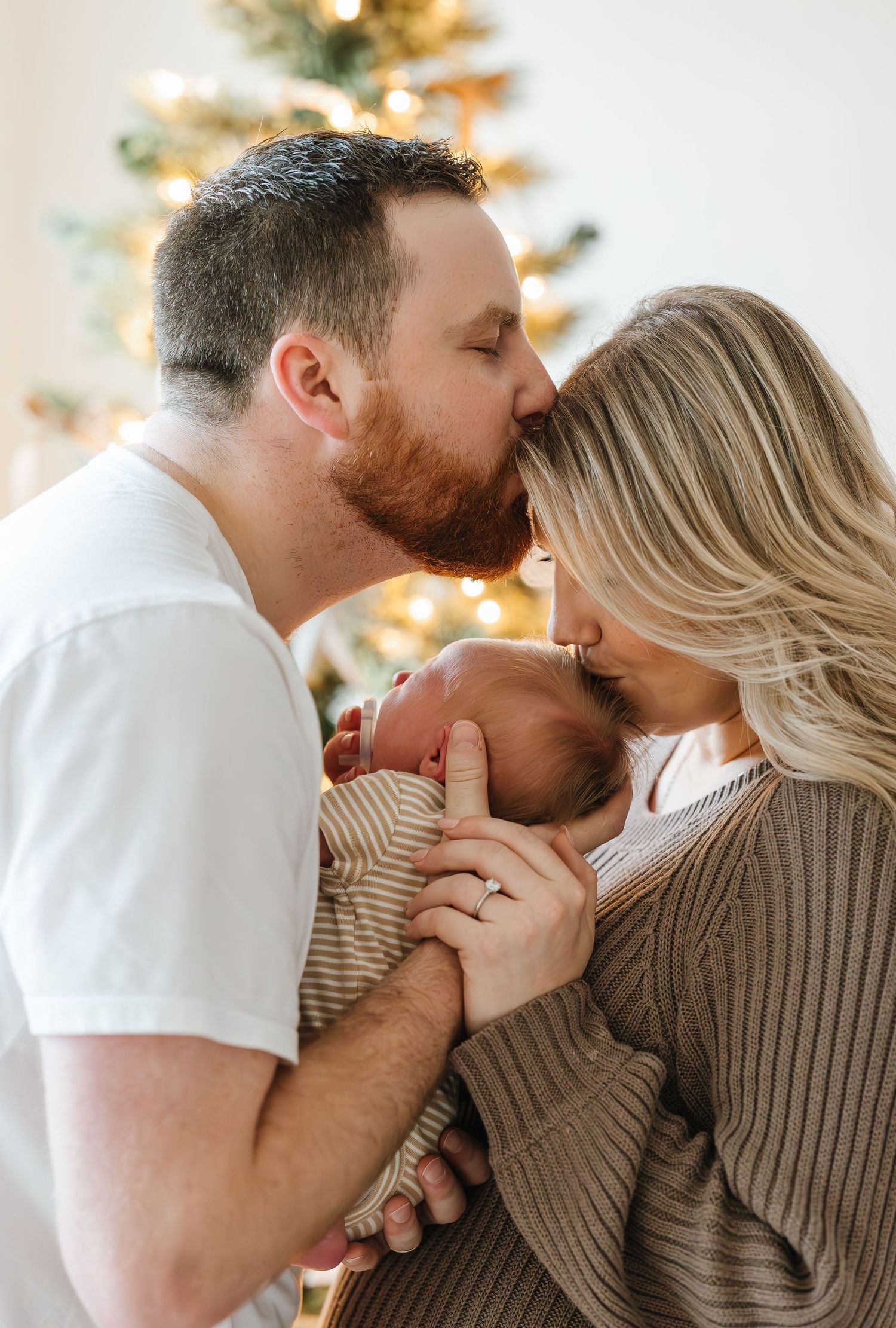 A tender family moment with a newborn baby near a Christmas tree in warm lighting.