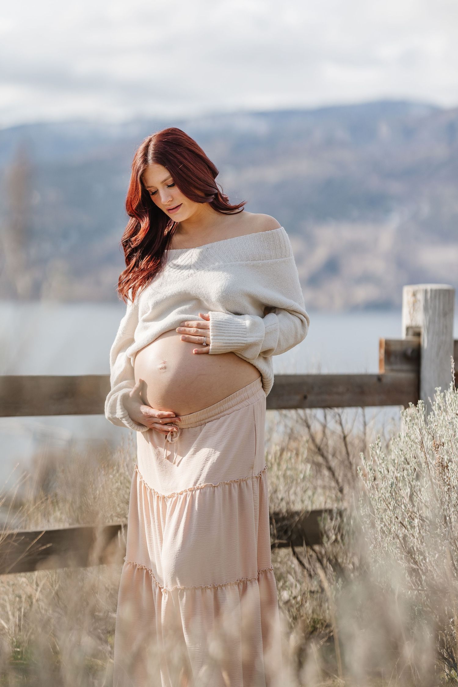 Maternity solo session with mother cradling belly while standing by rustic fence with mountain view at Knox mountain park in Kelowna
