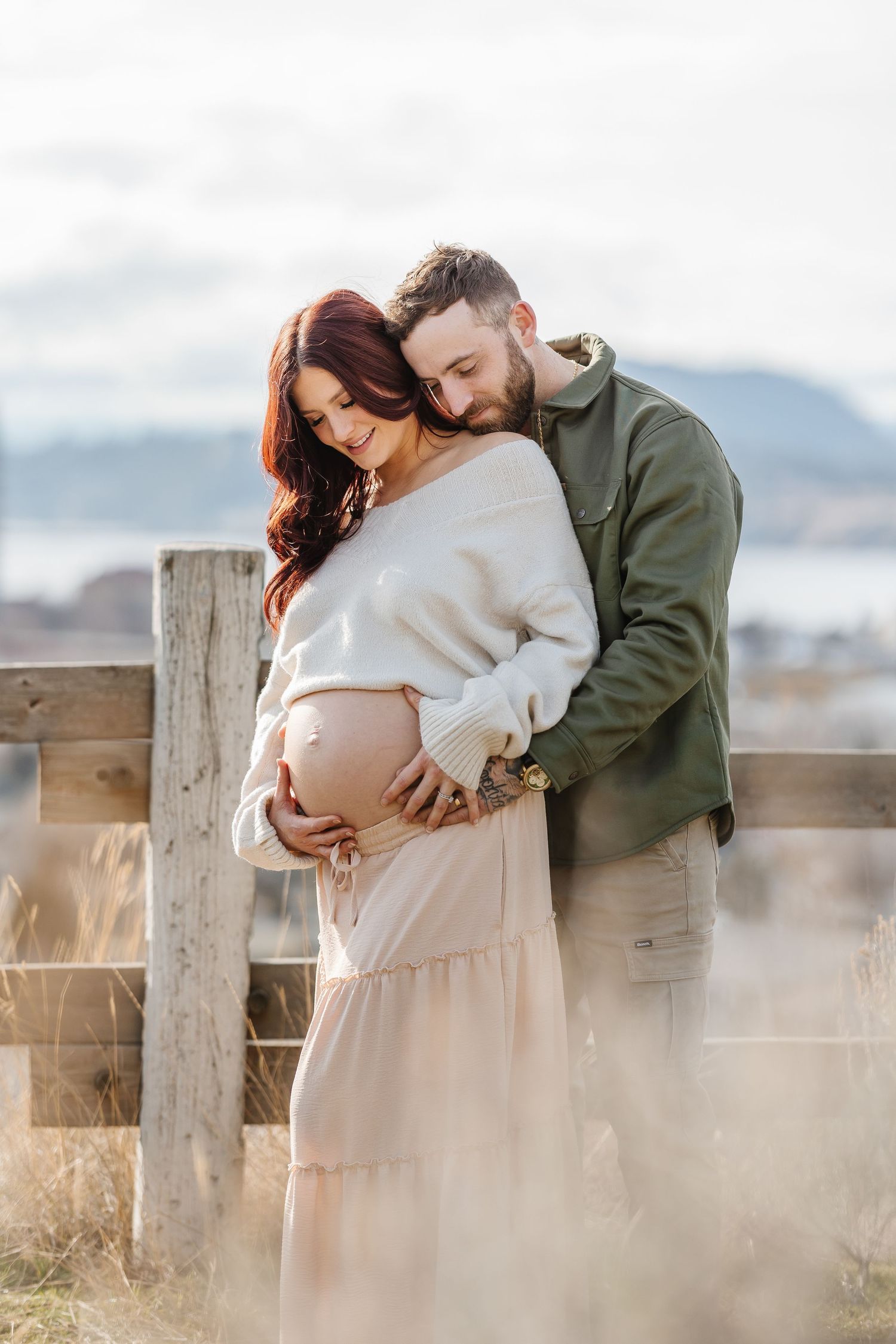 Couple embraces in a romantic maternity photo outdoors with mountains and wooden fence in the background at Knox mountain park in Kelowna