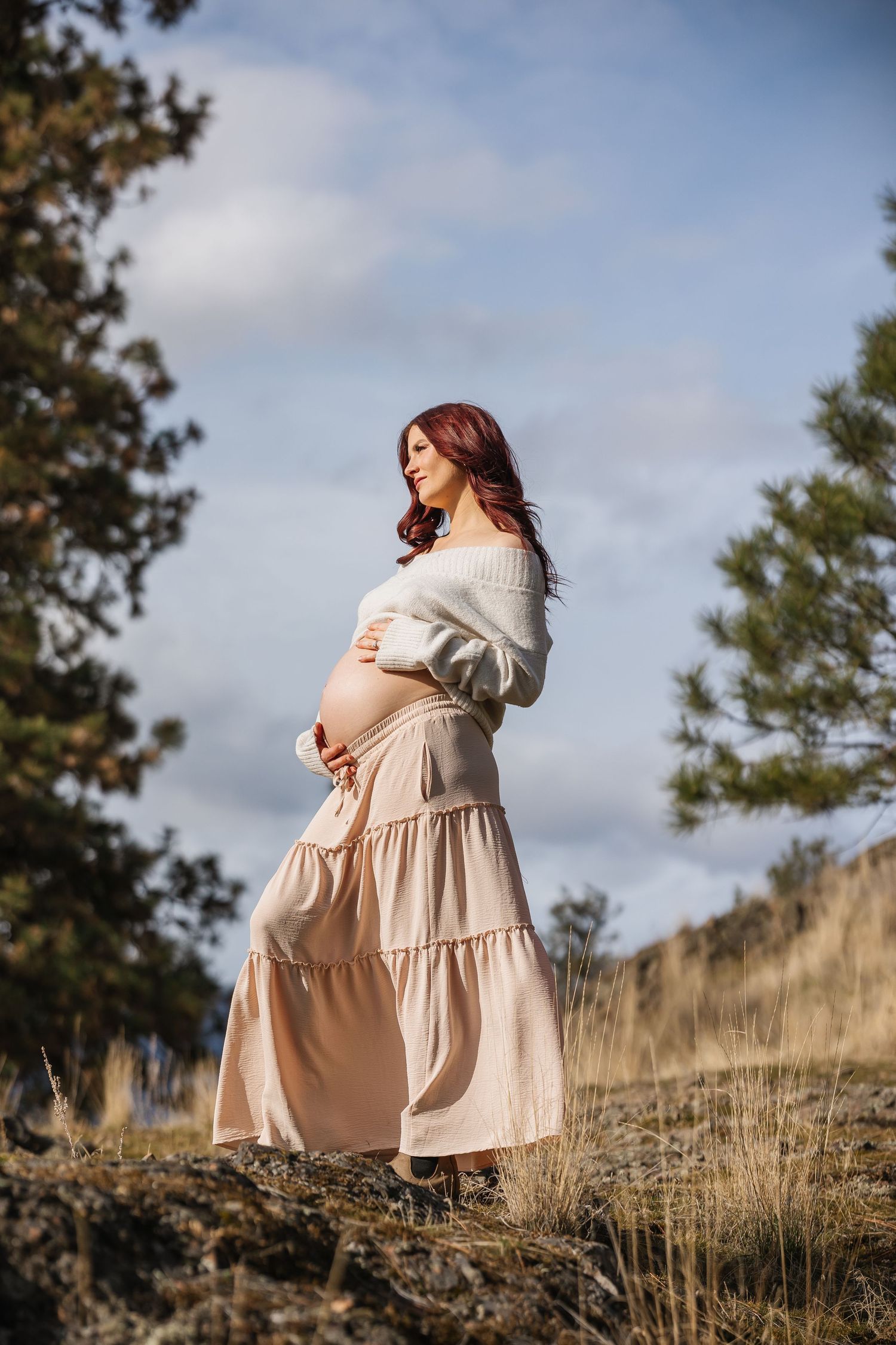 solo Maternity photo's in a flowing beige dress on a grassy hillside against a cloudy sky in a series of artistic photos in Kelowna bc
