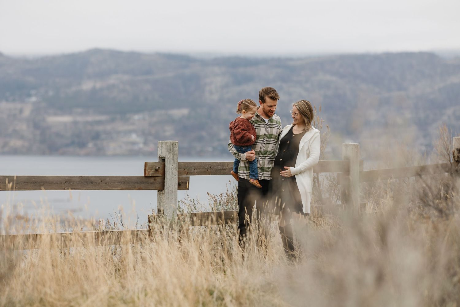 family photos sharing a candid moment looking at baby belly in a grassy field overlooking okanagan lake at Knox mountain park