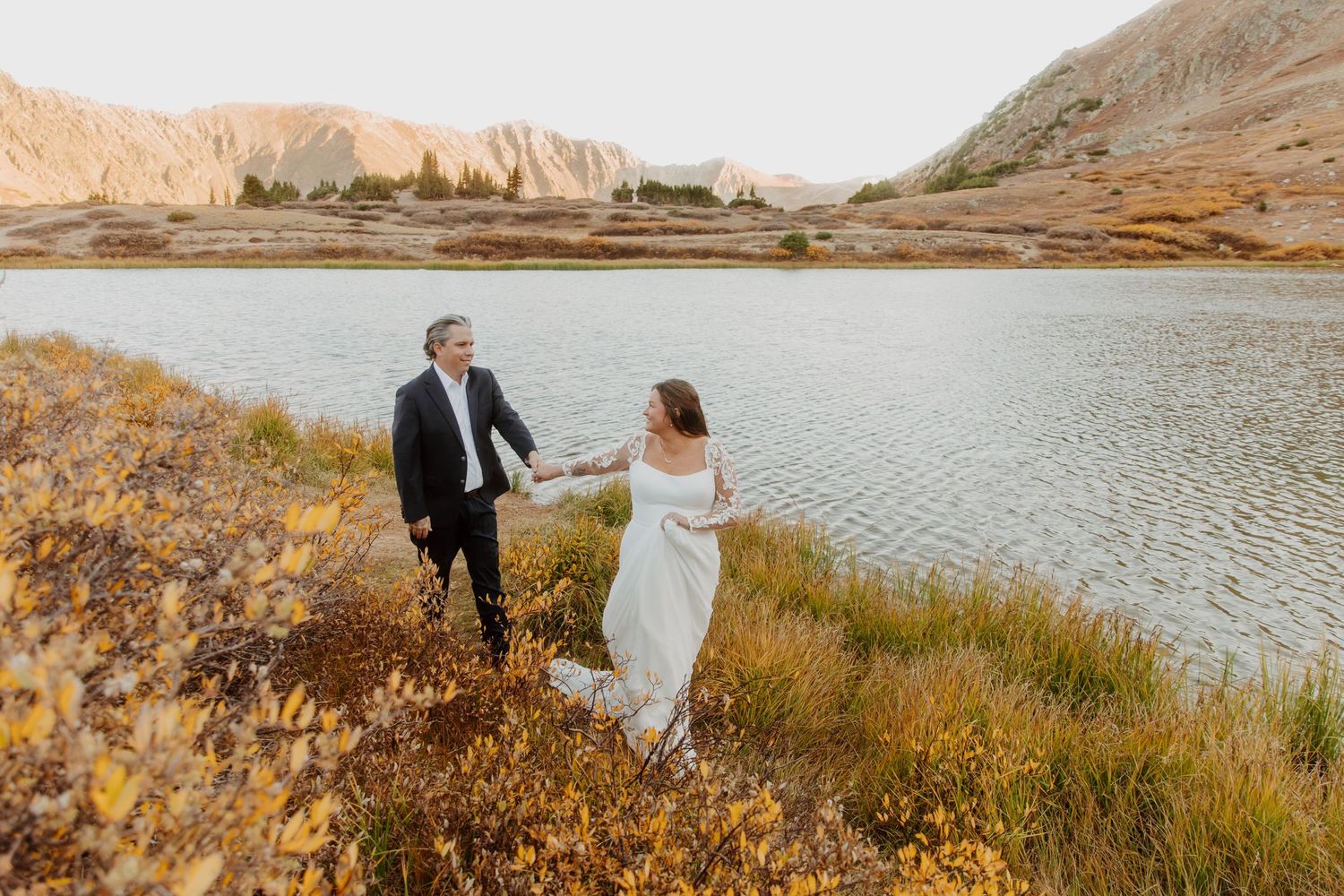 A couple in wedding attire holds hands by a serene lake surrounded by golden hills at sunset.