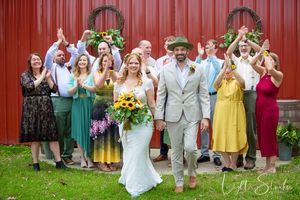 Wedding party celebrates with newlyweds in front of red barn with sunflowers and colorful summer attire.