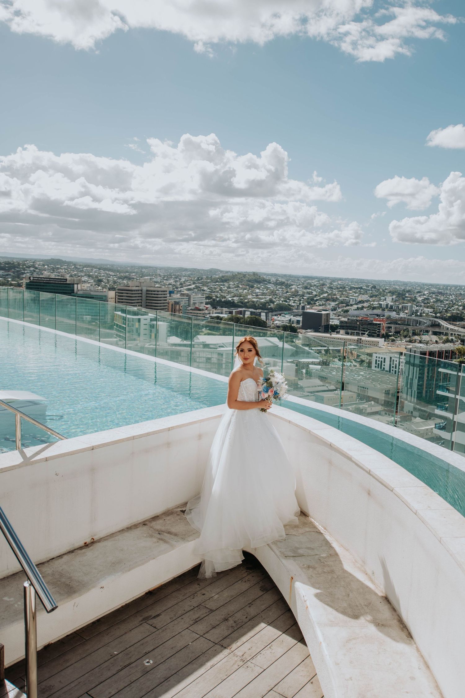 A bride in a white gown poses on a rooftop terrace with city views and infinity pool.