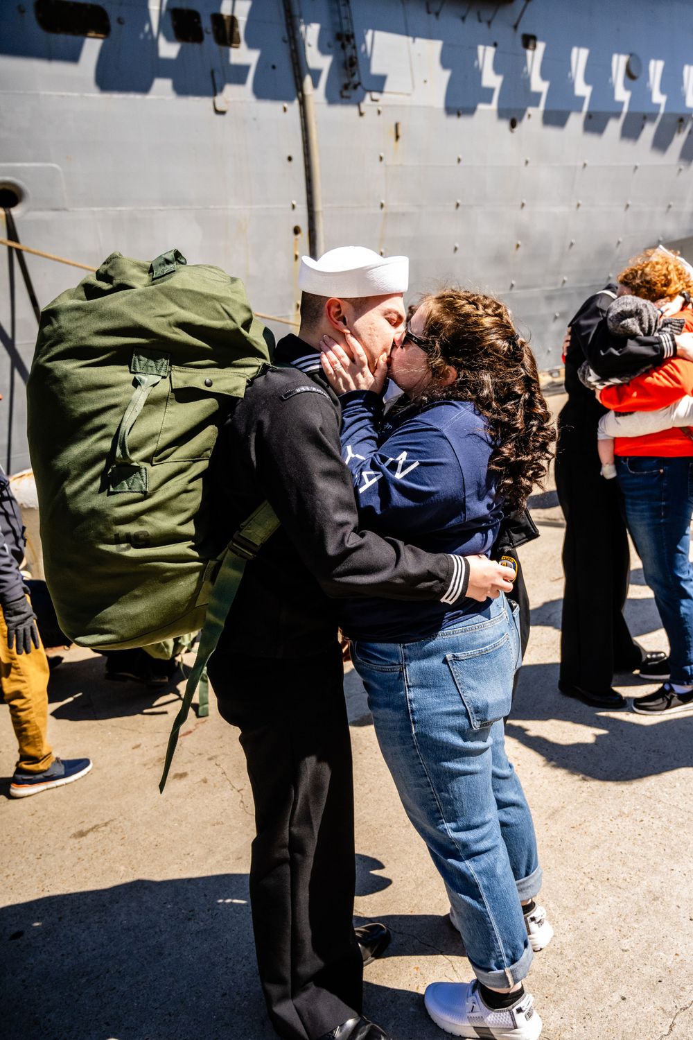 Military homecoming embrace between sailor and loved one beside naval vessel.