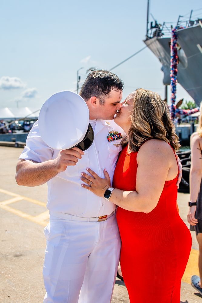Romantic reunion moment at naval base with ship visible in background.