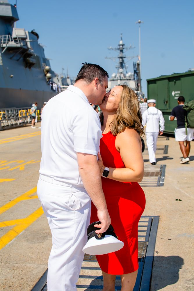 Emotional homecoming kiss at naval base with military ships in background.
