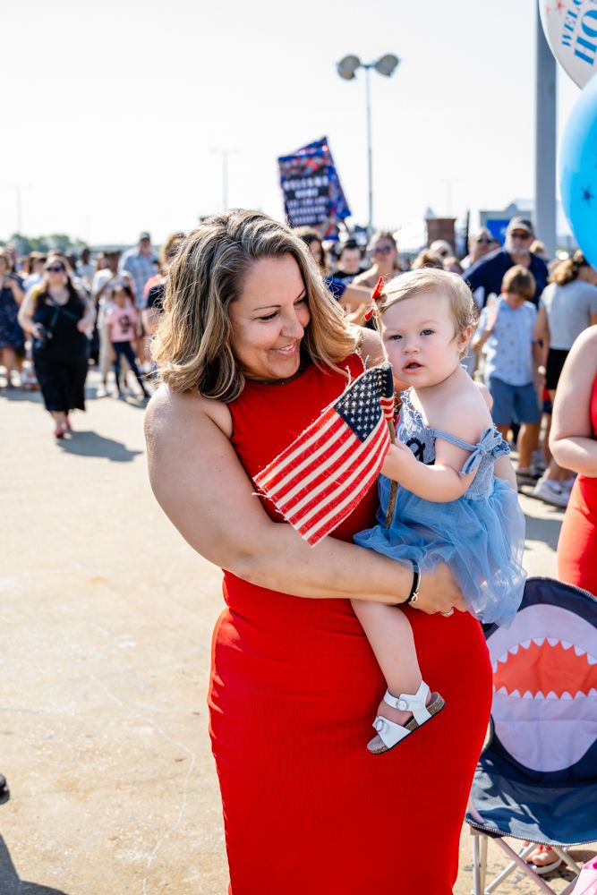 A festive outdoor gathering with a woman in a red dress holding a small child wearing blue against an American flag backdrop.