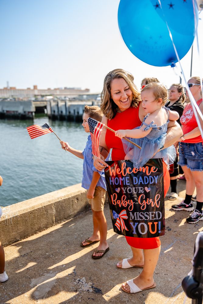 A waterfront homecoming celebration features decorative balloons and a welcome sign for a returning military member.