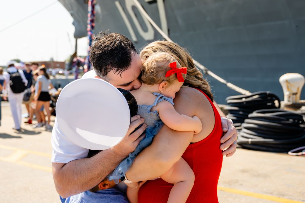 An emotional family reunion takes place outdoors near a large naval ship.