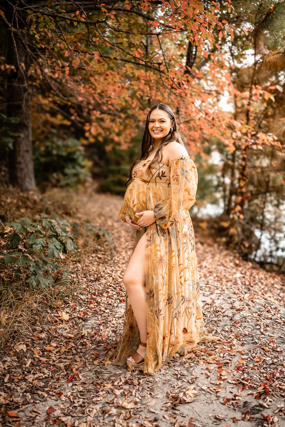 A maternity photo showcasing an elegant tan dress amidst autumn leaves by a river.
