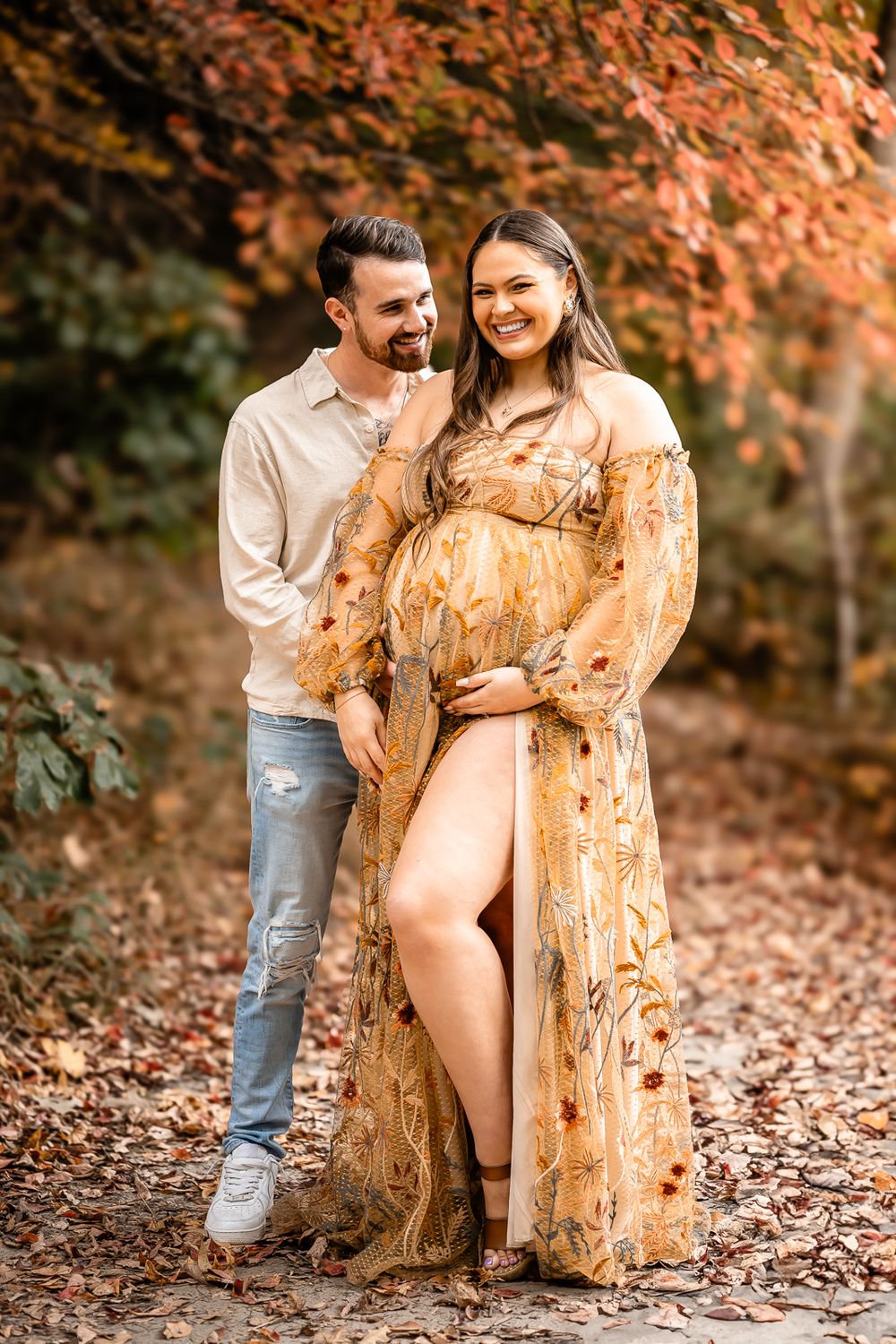 A romantic maternity photoshoot in autumn features a flowing golden dress against vibrant fall foliage.