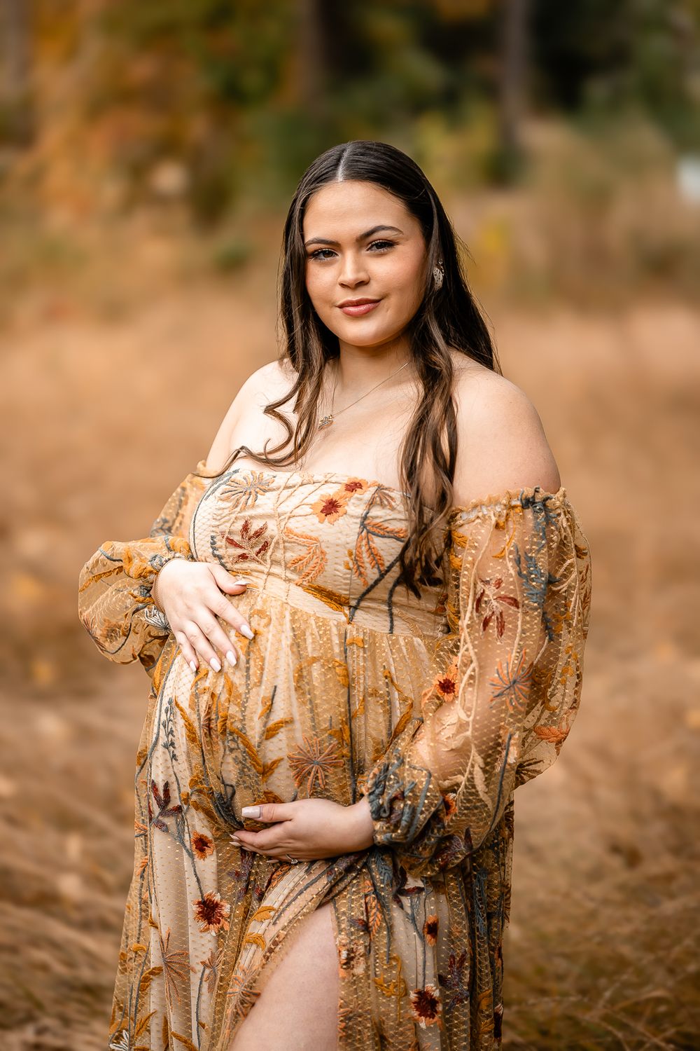 Expectant mother in a floral golden dress poses outdoors during fall season.