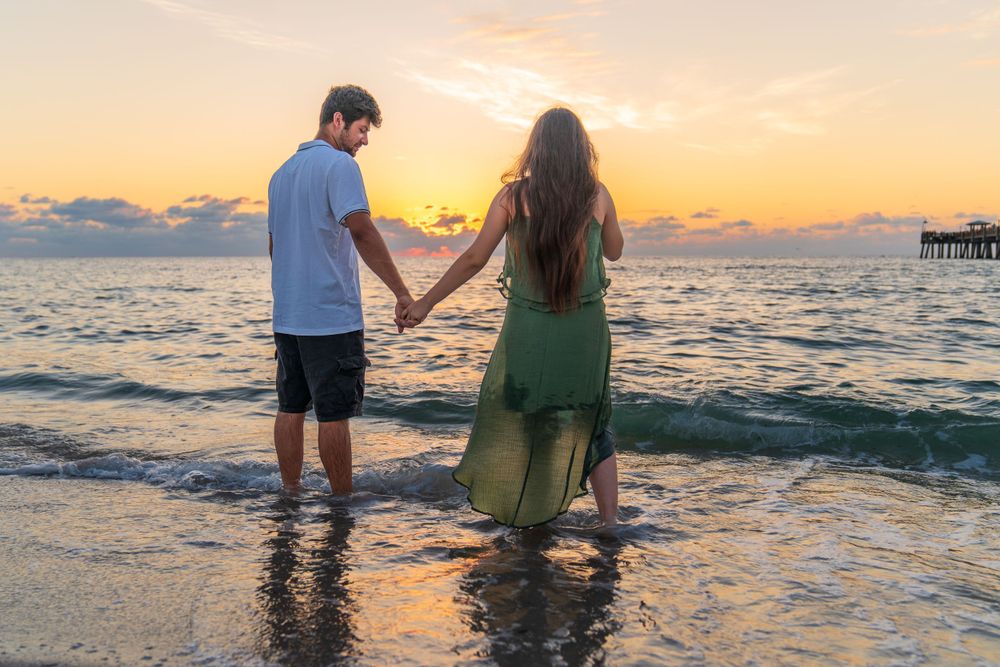 Couple holding baby bump during sunrise beach maternity session, blue skies