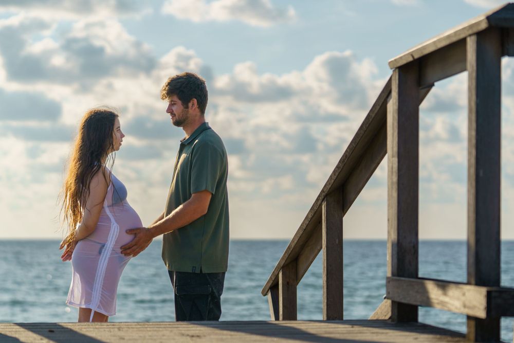 Couple holding baby bump during sunrise beach maternity session, blue skies