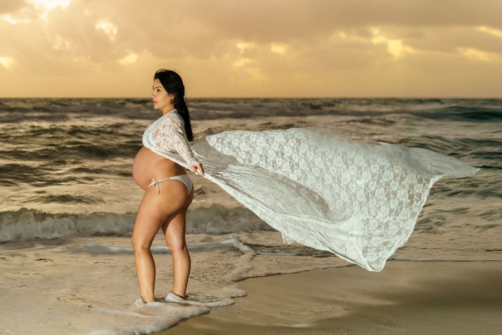 Pregnant mom in a flowing dress walking along the shoreline at sunset, relaxed beach maternity portrait by SRX Media Group.
