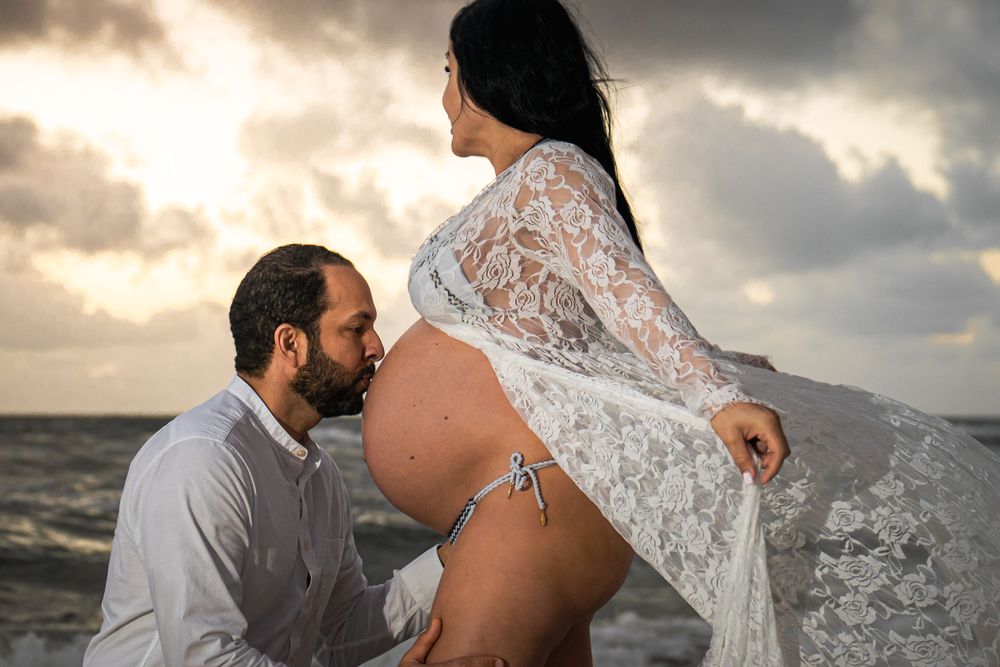 Couple holding baby bump during sunrise beach maternity session, golden hour, stormy skies