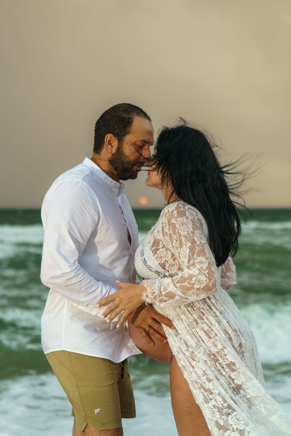 Couple holding baby bump during sunrise beach maternity session, golden hour, stormy skies