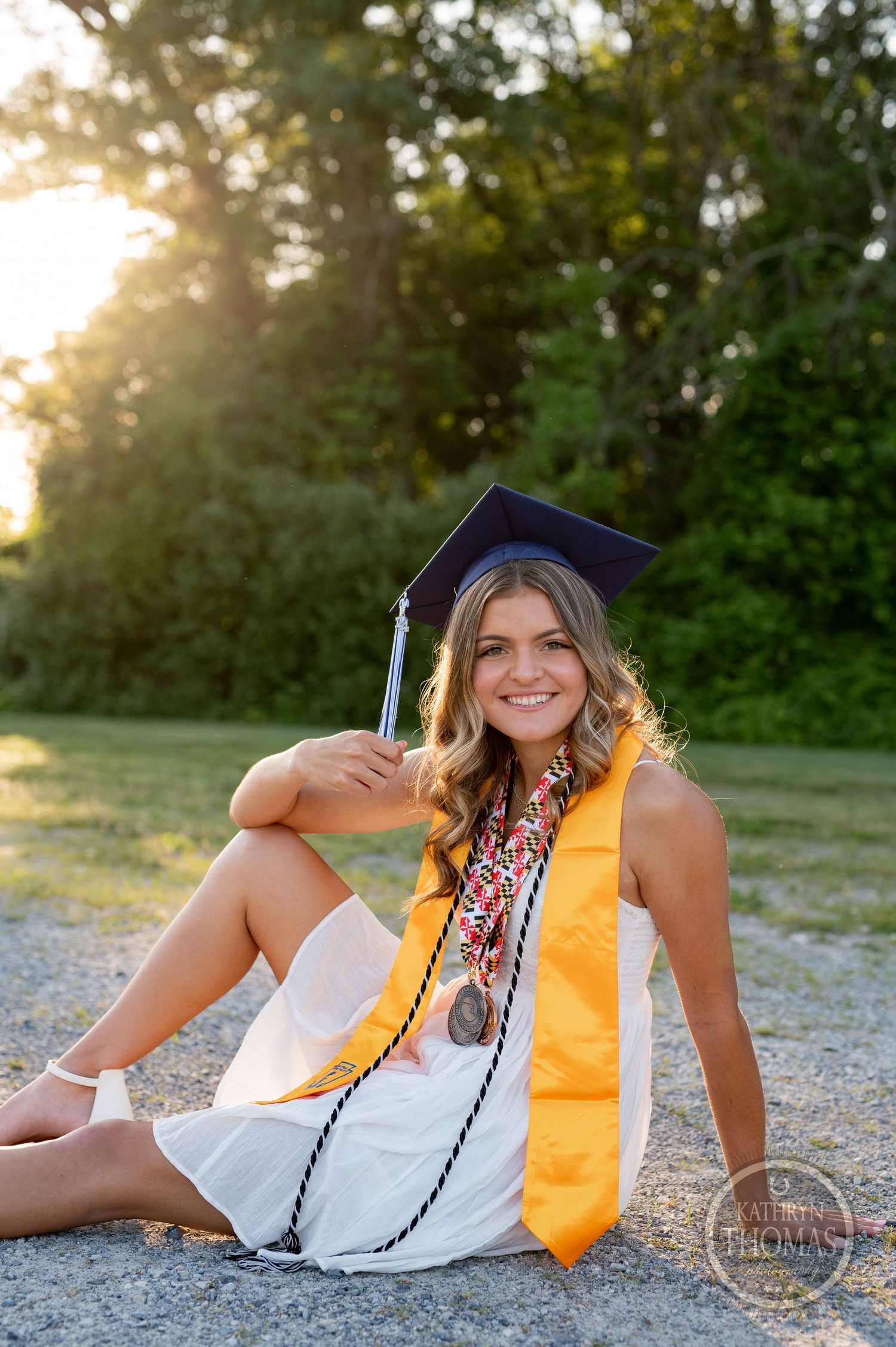 Graduate in white dress with yellow honors stole poses during golden hour photography session.