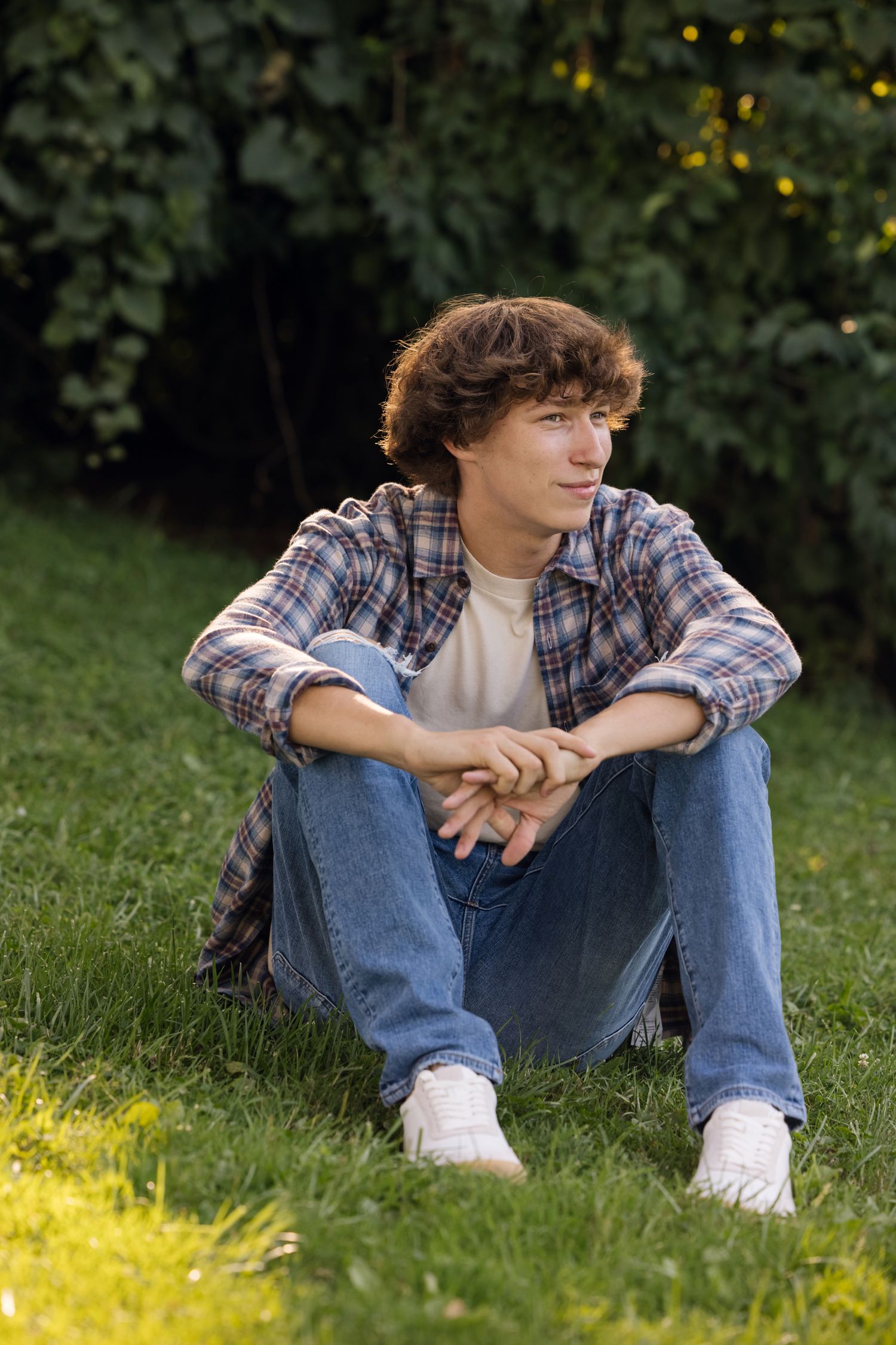 A casual outdoor portrait of someone sitting on grass wearing plaid and jeans in a garden setting.