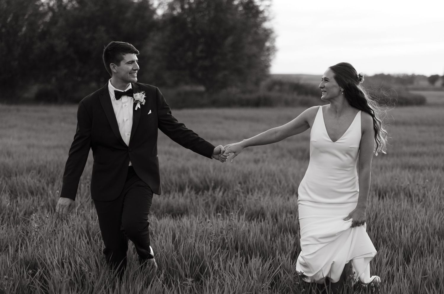 A black and white wedding photo of a couple holding hands while walking through a scenic field at dusk.