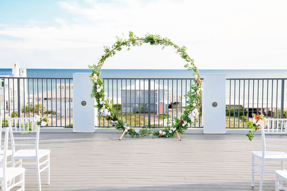 A seaside wedding ceremony setup with floral arch and white chairs on an oceanfront deck.