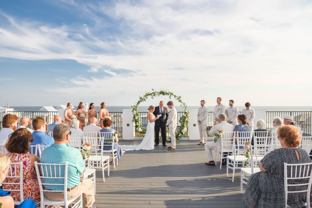 An outdoor beach wedding ceremony with guests seated under a floral arch.