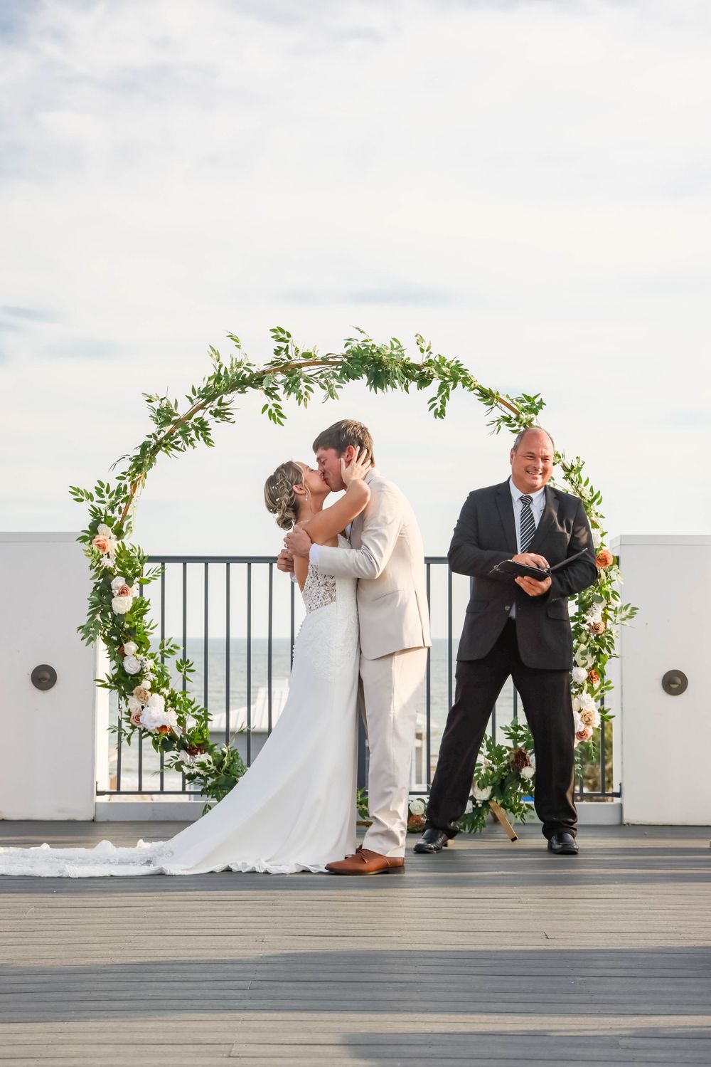A wedding ceremony kiss under a greenery arch with officiant standing nearby.