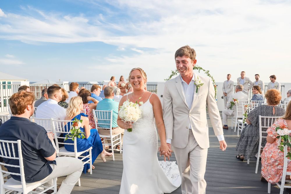 Newlyweds walking down the aisle while guests celebrate on a waterfront deck.