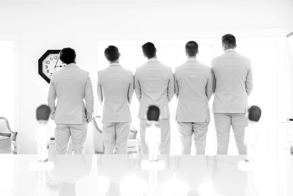 Groomsmen in white suits photographed from behind in artistic black and white style.