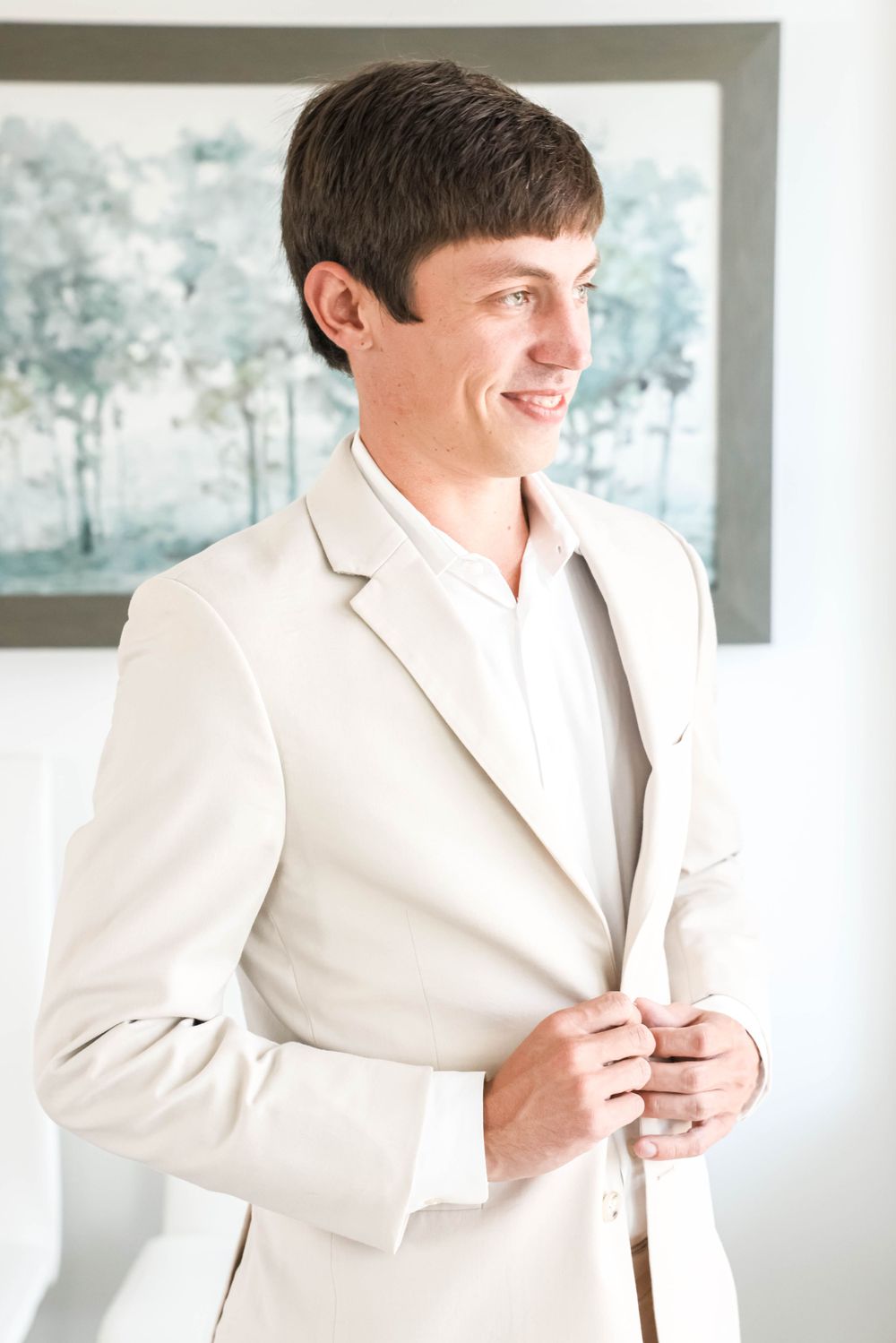 Groom in light beige suit smiles while getting ready in bright modern room.